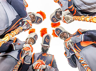 Members of the Marching Pride Band are looking down at the camera in a circle with their instruments