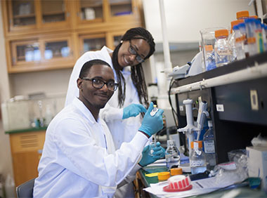 Inside a science lab, a male and female student are working and smiling at camera
