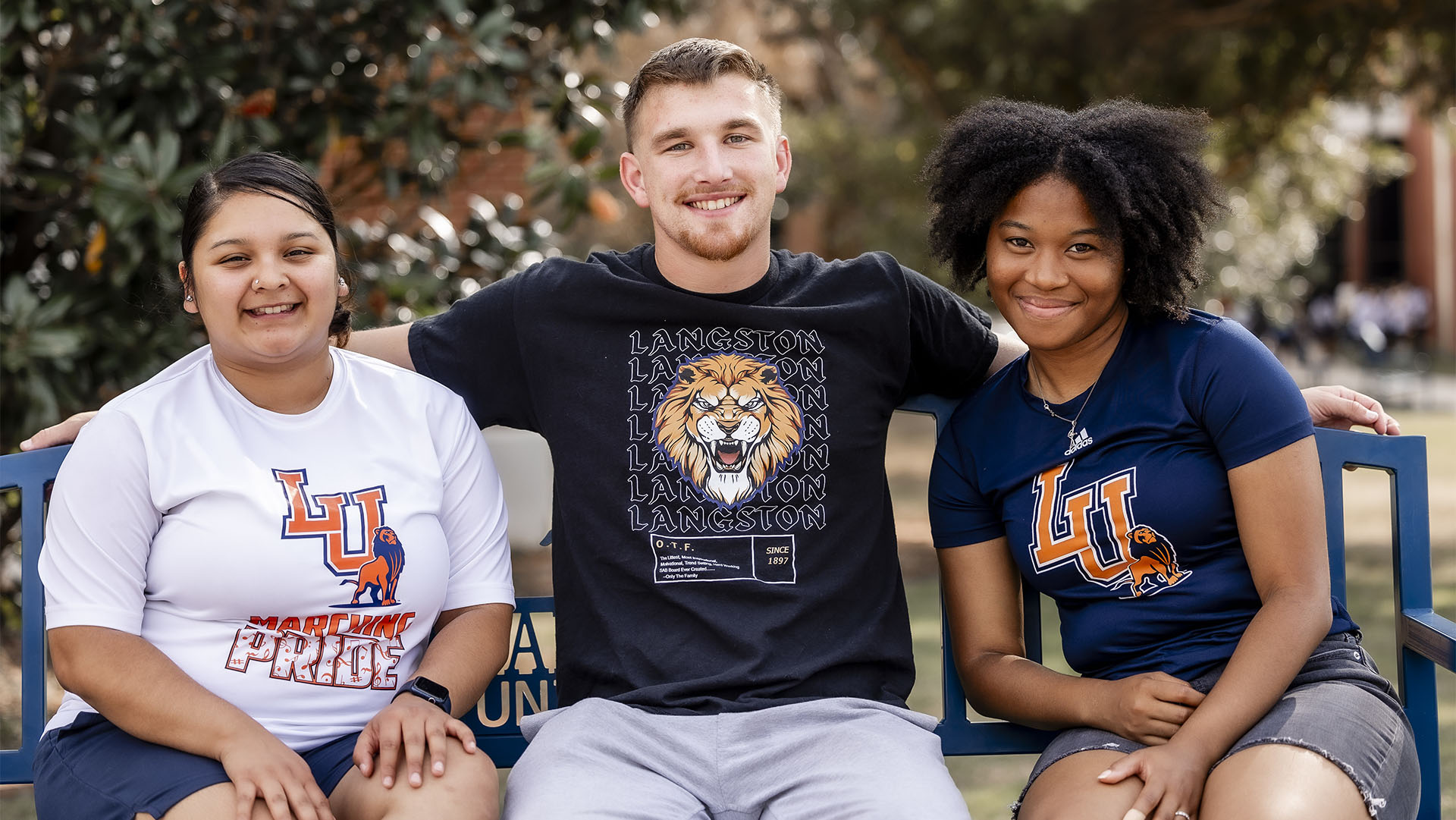 Photo of three students sitting on bench smiling facing forward on campus