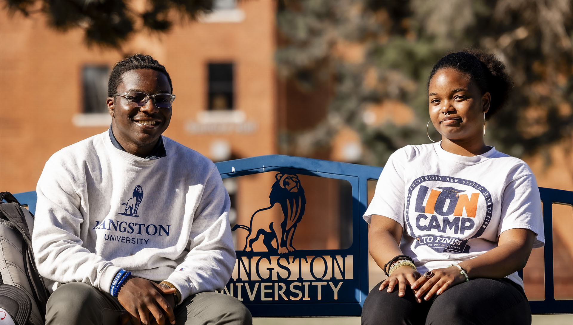 A male and female student in Langston University logo wear sitting on a bench smiling at the camera
