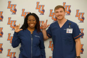 Soli Pannell (left) and Asher Bellavigna flash the L's Up while wearing their nursing scrubs and standing in front of an LU backdrop.