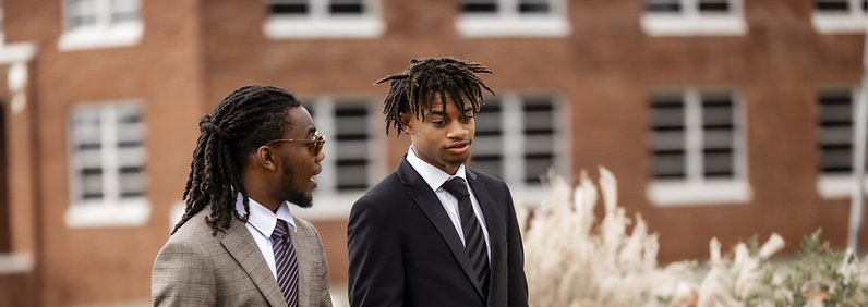 Two Langston University male students in business attire walking campus grounds and talking.