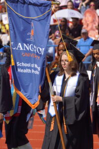 Kaitlyn Mitchell in graduation regalia walking with the School of Nursing and Health Professions' flag